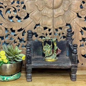 May include: A small wooden bench with carved details, featuring a brass statue of a couple on a swing. The bench is sitting on a wooden surface with intricate carvings.