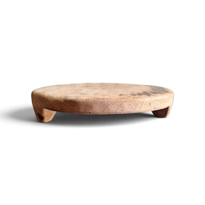 May include: A round, light brown wooden riser with a weathered, textured surface and visible grain patterns. The circular board is supported by three short, sturdy wooden feet, elevating it slightly above the surface it rests upon.