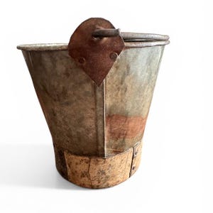 May include: An antique metal bucket with a weathered appearance. The bucket has a trapezoidal shape with a riveted wooden base and a rusty metal handle. The metal has a mottled gray and brown color, suggesting age and use. The bucket is likely for decorative purposes.