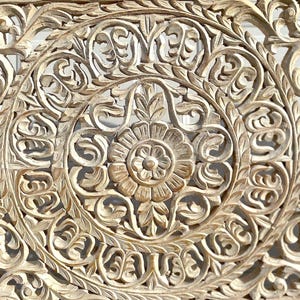 May include: Intricately carved wooden wall decor with a whitewashed finish. The design features a central floral motif surrounded by swirling patterns.
