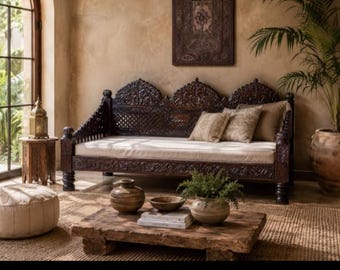 May include: A dark brown carved wooden daybed with a cream-colored cushion and three decorative pillows. The frame features intricate lattice and floral carvings, complemented by a low wooden coffee table and a round beige floor pouf.