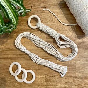 Peut inclure: Un ensemble de trois anneaux en bois et un porte-plante en macramé avec un anneau en bois en haut. Le porte-plante est fait de corde de coton naturelle et est de couleur beige clair. Les anneaux en bois sont également de couleur beige clair.