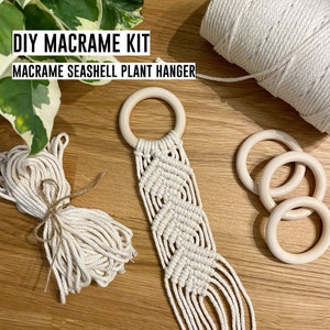 May include: A DIY macrame kit for a seashell plant hanger. The kit contains off-white macrame cord, wooden rings, and a completed macrame plant hanger. The image text reads "DIY MACRAME KIT" and "MACRAME SEASHELL PLANT HANGER."