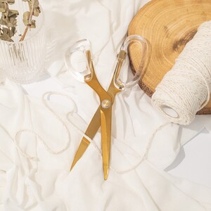 Può includere: Forbici dorate con manici in plastica trasparente su uno sfondo di tessuto bianco. È visibile anche un rotolo di spago.