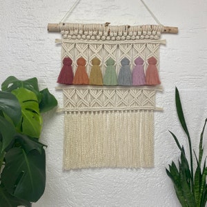 May include: A macrame wall hanging with a white cotton cord and rainbow coloured tassels. The wall hanging is made with a knotting technique and has a fringe at the bottom.
