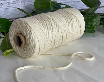 Könnte beinhalten: Eine Spule aus natürlichem weißem Baumwollseil mit einem Stück Seil, das von der Spule abgeht. Das Seil liegt auf einer weißen Oberfläche mit grünen, blättrigen Pflanzen im Hintergrund.