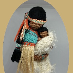 Puede incluir: Muñeca de crochet de una mujer nativa americana sosteniendo un bebé. La mujer lleva un vestido turquesa, naranja y marrón con flecos blancos. El bebé está envuelto en una manta blanca. La muñeca está hecha por Cubby House Crochet.