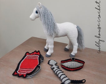 May include: A white crocheted horse with a gray mane and tail. The horse is wearing a red and white saddle, a gray and black bridle, and a gray and black stirrup.