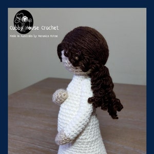 May include: Crocheted doll of a pregnant woman with brown hair, wearing a white dress. The doll is standing on a wooden surface. The text "Mother" and "Proverbs 31:25" are written below the doll. The text "Crochet Pattern" is written below the text.