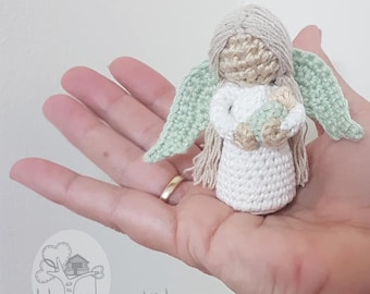 May include: A crocheted angel figurine holding a smaller crocheted angel in its arms. The angel has long blonde hair, a white dress, and green wings. The smaller angel is also white with a green halo.