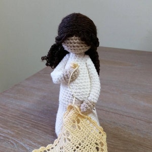 May include: A crocheted doll with brown hair is holding a crochet hook and yarn. The doll is wearing a white dress and is crocheting a yellow doily. The doll is sitting on a wooden table. The text "Cubby House Crochet" is visible in the image.