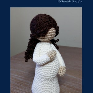 May include: Crocheted doll of a pregnant woman with brown hair. The doll is wearing a white dress and has a light brown complexion. The doll is standing on a wooden surface. The text "Mother" is written in cursive above the doll. The text "Proverbs 31:25" is written below the doll. The text "Cubby House Crochet" is written below the doll. The text "Made in Australia by Veronica McRae" is written below the doll.