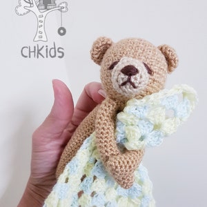 Puede incluir: Manta de seguridad de oso de peluche de ganchillo en color beige, crema y azul claro. El oso tiene un cuerpo beige, cara crema y ojos cerrados. La manta es un patrón de tejido abierto suave en azul claro y crema. Incluye el texto "CHKids" y un logotipo de casa en el árbol.