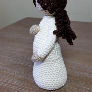 May include: Crocheted doll with brown hair in a white dress. The doll is pregnant and has a baby bump. The doll is standing on a wooden surface. The text "Mother" is written below the doll, with the words "Proverbs 31:25" underneath.