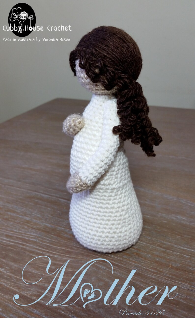 May include: Crocheted doll with brown hair in a white dress. The doll is pregnant and has a baby bump. The doll is standing on a wooden surface. The text "Mother" is written below the doll, with the words "Proverbs 31:25" underneath.