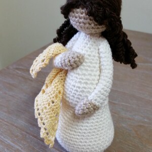 May include: A crocheted doll with brown hair, wearing a white dress and holding a yellow crocheted shawl. The doll is standing on a wooden surface. The text "Cubby House Crochet" and "Made in Australia by Veronica McRae" is visible below the doll.