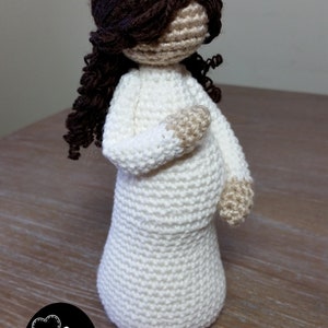 May include: A crocheted doll with long brown hair wearing a white dress. The doll is standing on a wooden surface. The text "Cubby House Crochet" is visible at the bottom of the image.