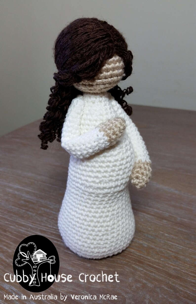 May include: A crocheted doll with long brown hair wearing a white dress. The doll is standing on a wooden surface. The text "Cubby House Crochet" is visible at the bottom of the image.