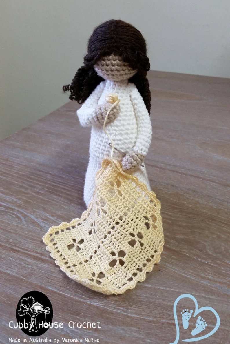 May include: A crocheted doll with brown hair is holding a crochet hook and yarn. The doll is wearing a white dress and is crocheting a yellow doily. The doll is sitting on a wooden table. The text "Cubby House Crochet" is visible in the image.
