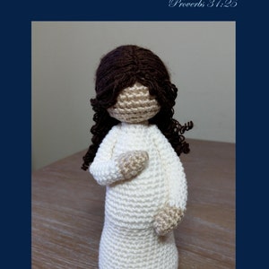 May include: Crocheted doll with brown hair and a white dress. The doll is standing on a wooden surface. The text "Mother" is written in cursive above the doll. The text "Proverbs 31:25" is written below the doll. The text "Cubby House Crochet" is written below the doll. The text "Made in Australia by Veronica McRae" is written below the doll.