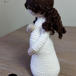 May include: Crocheted doll of a pregnant woman with brown hair, wearing a white dress. The doll is standing on a wooden surface. The text "Mother Proverbs 31:25" is written above the doll. The text "Cubby House Crochet Made in Australia by Veronica McRae" is written below the doll.