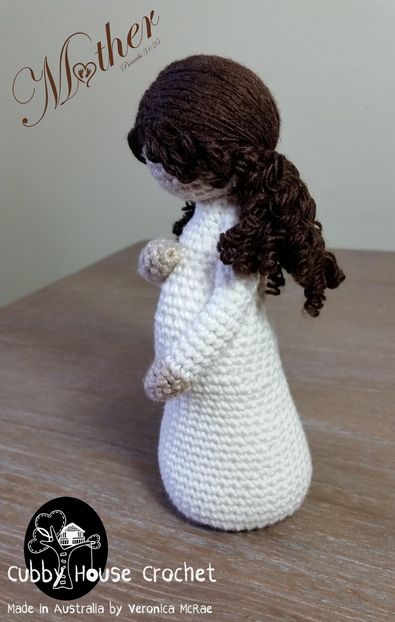 May include: Crocheted doll of a pregnant woman with brown hair, wearing a white dress. The doll is standing on a wooden surface. The text "Mother Proverbs 31:25" is written above the doll. The text "Cubby House Crochet Made in Australia by Veronica McRae" is written below the doll.