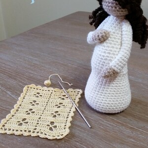 May include: A crocheted doll with brown hair, wearing a white dress, standing on a wooden table with a crocheted square and a crochet hook. The doll is pregnant and has a large belly. The text "Cubby House Crochet" and "Made in Australia by Veronica McRae" is visible at the bottom of the image.