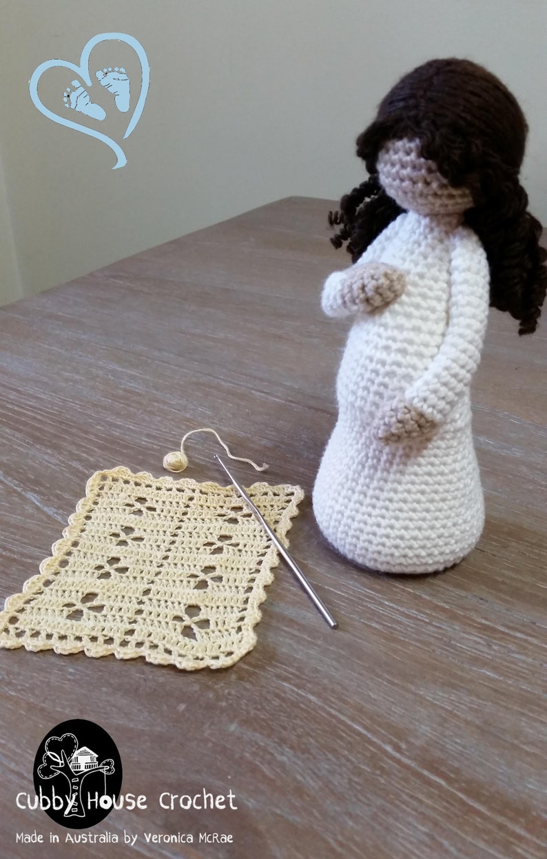 May include: A crocheted doll with brown hair, wearing a white dress, standing on a wooden table with a crocheted square and a crochet hook. The doll is pregnant and has a large belly. The text "Cubby House Crochet" and "Made in Australia by Veronica McRae" is visible at the bottom of the image.