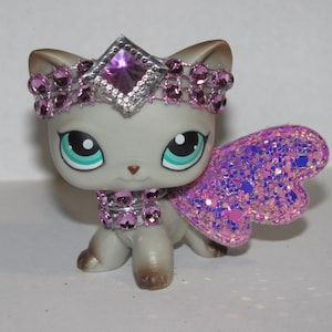 Pu&ograve; includere: Statuina di gatto grigio con grandi occhi turchese, ornata da una scintillante corona rosa, collare e ali. Le ali sono un mix di glitter rosa e viola. La corona presenta una grande gemma viola a forma di diamante.