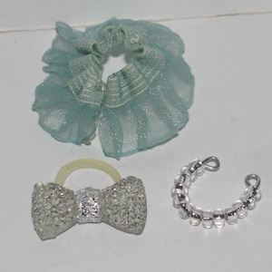 Puede incluir: Tres accesorios plateados para una mu&ntilde;eca o un animal peque&ntilde;o. Un lazo plateado con una banda el&aacute;stica transparente, un collar de cadena plateada y un collar de tul azul claro.