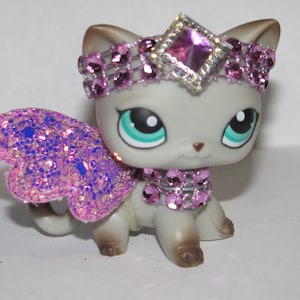 Pu&ograve; includere: Una statuina di gatto grigio con una tiara, un collare e ali glitterate rosa. Il gatto ha grandi occhi verde-azzurri e un piccolo naso rosa. Le ali sono un mix di glitter rosa e blu.