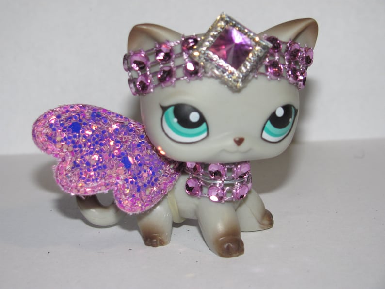 Pu&ograve; includere: Una statuina di gatto grigio con una tiara, un collare e ali glitterate rosa. Il gatto ha grandi occhi verde-azzurri e un piccolo naso rosa. Le ali sono un mix di glitter rosa e blu.