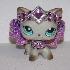 Pu&ograve; includere: Figurina di gatto grigio con grandi occhi blu-verdi, adornata da un diadema, collare e ali scintillanti viola e argento. Il gatto ha zampe e orecchie marroni, creando un aspetto stravagante e decorativo.