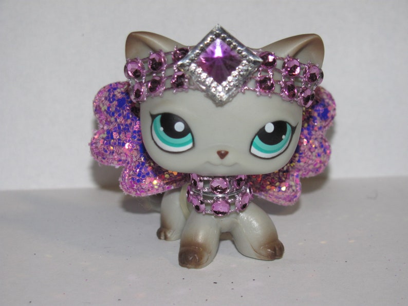 Pu&ograve; includere: Figurina di gatto grigio con grandi occhi blu-verdi, adornata da un diadema, collare e ali scintillanti viola e argento. Il gatto ha zampe e orecchie marroni, creando un aspetto stravagante e decorativo.