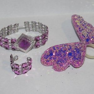 Pu&ograve; includere: Un set di bigiotteria tra cui un braccialetto, un anello e un accessorio per capelli a forma di ala. Il braccialetto e l'anello sono color argento con pietre preziose rosa. L'accessorio ad ala &egrave; rosa con glitter e accenti blu.