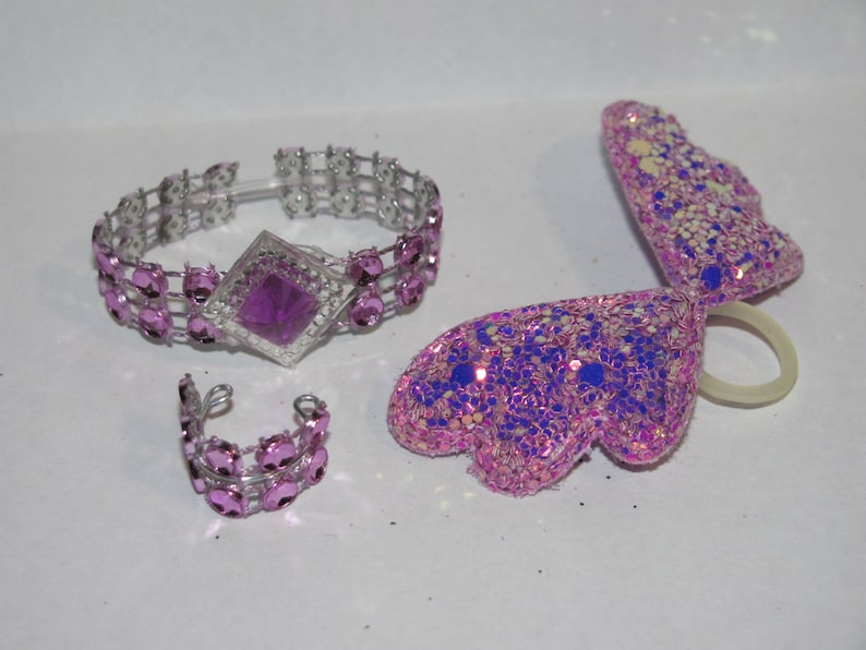 Pu&ograve; includere: Un set di bigiotteria tra cui un braccialetto, un anello e un accessorio per capelli a forma di ala. Il braccialetto e l'anello sono color argento con pietre preziose rosa. L'accessorio ad ala &egrave; rosa con glitter e accenti blu.