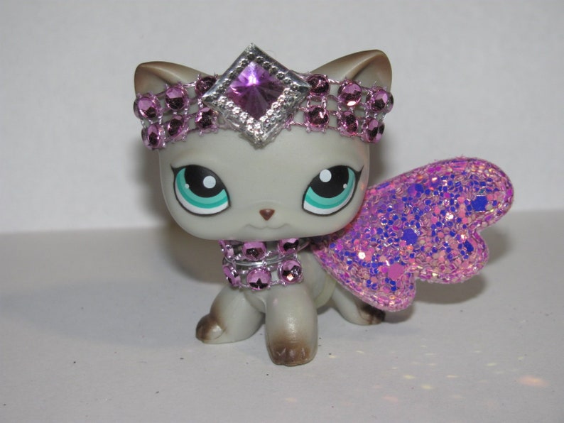 Pu&ograve; includere: Statuina di gatto grigio con grandi occhi turchese, ornata da una scintillante corona rosa, collare e ali. Le ali sono un mix di glitter rosa e viola. La corona presenta una grande gemma viola a forma di diamante.
