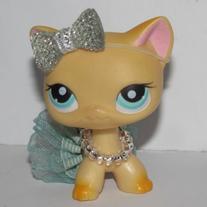 Puede incluir: Un peque&ntilde;o juguete de gato de color beige con un lazo de purpurina plateado, una falda de tul azul claro y un collar de cuentas plateadas.