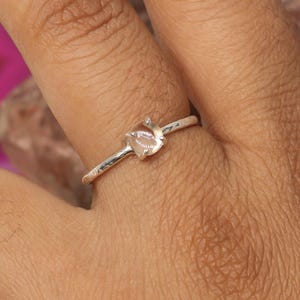 Peut inclure: Une bague en argent avec une petite pierre précieuse de couleur claire. La bague est portée au doigt, mettant en valeur la forme de la pierre et le corps de la bague. L'arrière-plan comprend des pierres polies colorées.