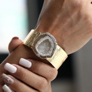 Puede incluir: Un brazalete dorado con un acabado texturizado. El brazalete presenta una piedra de geoda central con un interior cristalino blanco y gris. El brazalete se lleva en la muñeca.