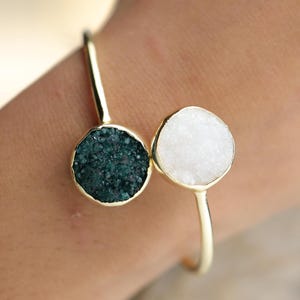 Könnte beinhalten: Ein goldfarbenes offenes Armband mit zwei kreisförmigen Fassungen. Eine Fassung enthält einen dunkelblau-grünen, texturierten Druzy-Stein, während die andere einen weißen, texturierten Druzy-Stein hält. Das Metallband ist glatt und poliert.