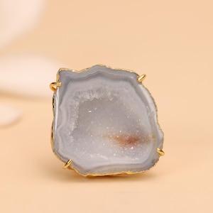 May include: A ring featuring a raw, irregular slice of light gray agate stone with a sparkling druzy center. The stone is encased in a thin, polished gold-tone metal setting with four small prongs securing the edges of the mineral specimen.