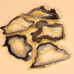 May include: A collection of five irregular agate slices featuring dark brown and grey banded patterns with central crystalline geode cavities. Each slice is framed by a metallic gold electroplated edge and includes small gold jump ring loops.
