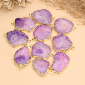 May include: A collection of irregular, flat purple agate slices with gold-toned metal edges. Each stone features a unique translucent crystalline pattern and includes small metal loops on the sides for connecting, creating a set of jewelry components.