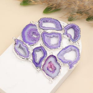 May include: A collection of irregular purple agate geode slices with silver-toned metallic edges. Each piece features a central hollow cavity surrounded by concentric purple and white crystalline bands, with small metal loops attached for jewelry making.