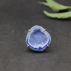 May include: A blue geode ring with gold-coloured prongs. The geode has a dark blue centre with lighter blue and white banding around the edges. The ring is set against a dark background.