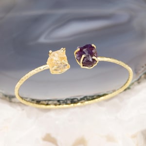 May include: Gold-tone open cuff bracelet featuring a rough-cut citrine and amethyst gemstone. The bracelet has a hammered texture and a minimalist design, perfect for everyday wear. The gemstones are a light yellow and deep purple color.