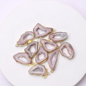May include: A collection of ten geode slices with a gold-colored trim and a small loop for hanging. The geodes have a light purple and white color scheme. These natural stone pendants are ideal for jewelry making.