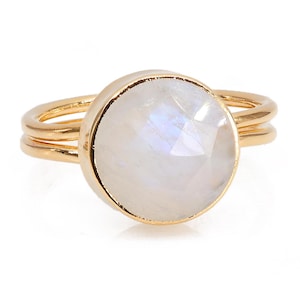 Pode incluir: Um anel dourado com uma grande pedra da lua redonda e iridescente. A pedra da lua tem uma superfície facetada que capta a luz. O anel tem um design de banda dupla.