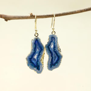 Puede incluir: Un par de pendientes colgantes dorados con piedras de ágata azul. Las piedras tienen una forma natural e irregular con un borde dorado. Los pendientes cuelgan de una fina rama marrón sobre un fondo claro.